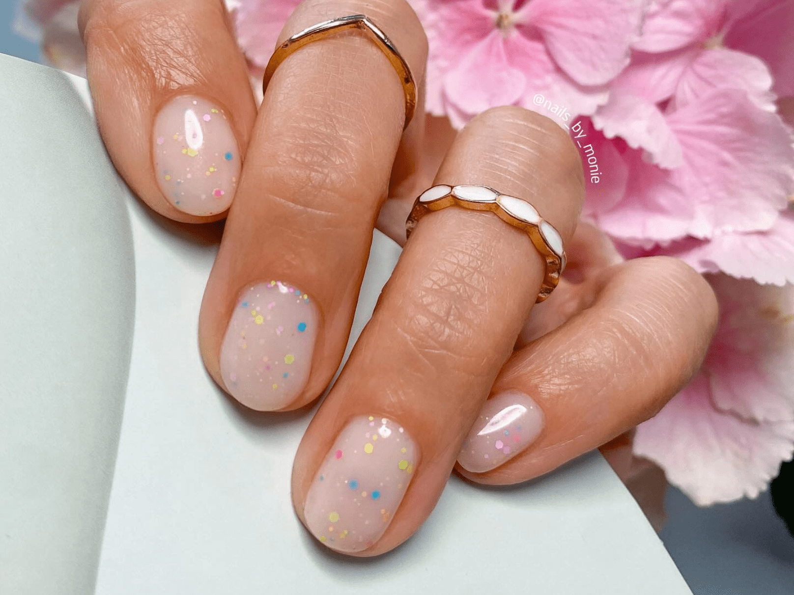 Hand holding pastel pink nail polish full of colorful dots, decorated with rings, against a background of pink flowers.