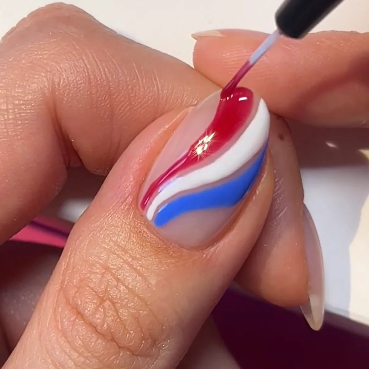 A close-up of a hand decorating a nail with wavy lines in red, white and blue.