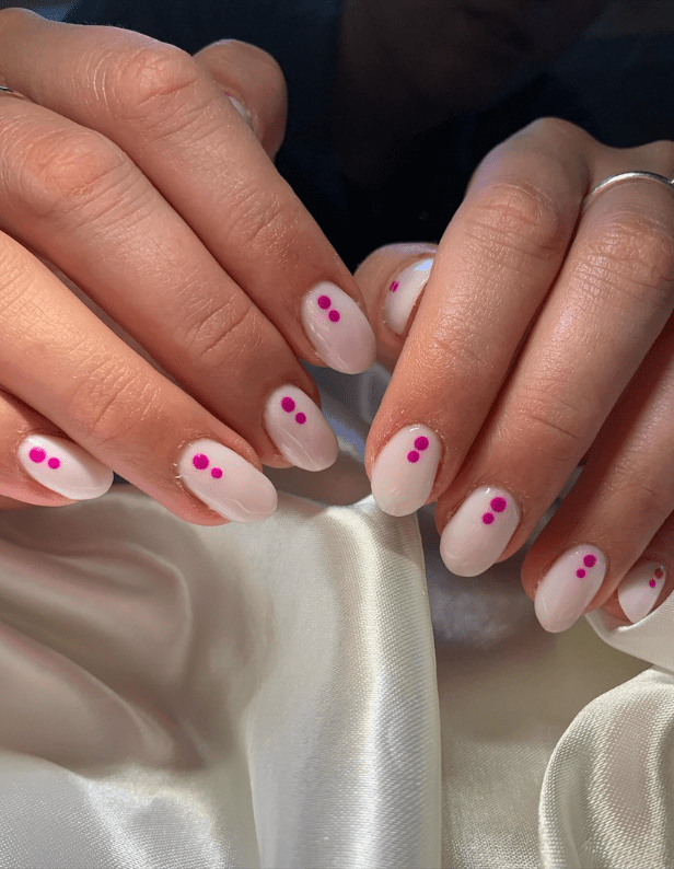 Two hands with light pink nail polish and fuchsia dots, resting on a white satin fabric.