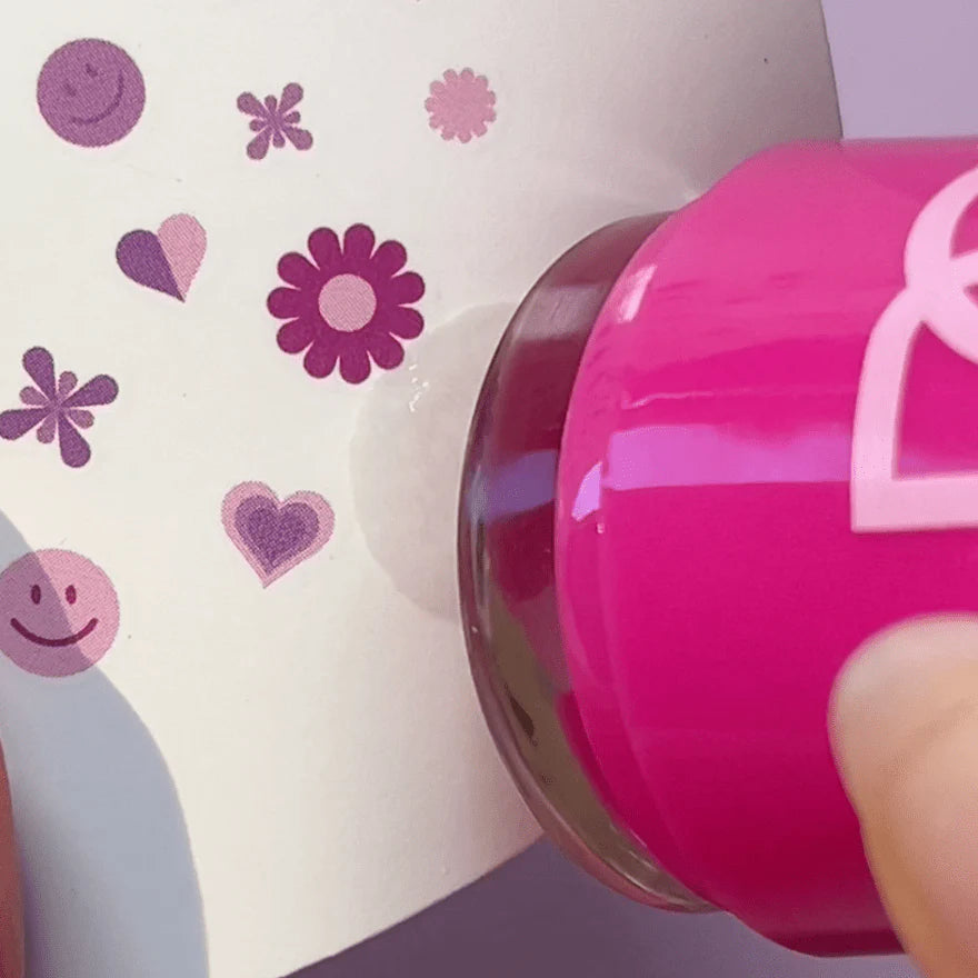 A pink stamp produces an imprint with purple and pink symbols such as flowers, hearts, and smiley faces on paper.