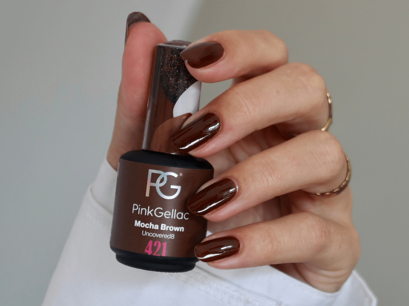 A hand with manicured nails, painted in a brown color, holds a bottle of brown nail polish.