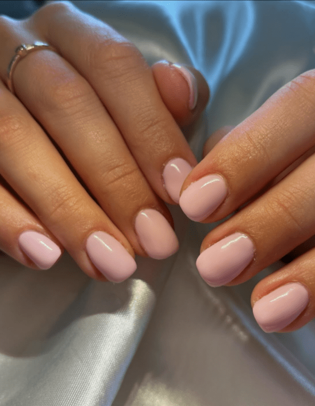 Two hands with short-painted light pink nails on a silk surface.