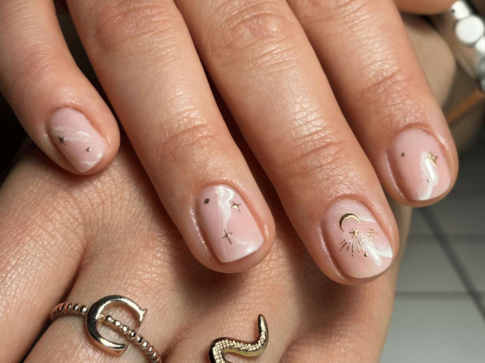 Close-up of a hand with nails painted in a light pink color, decorated with gold moon and star designs, with snake ring and buckle on the fingers.