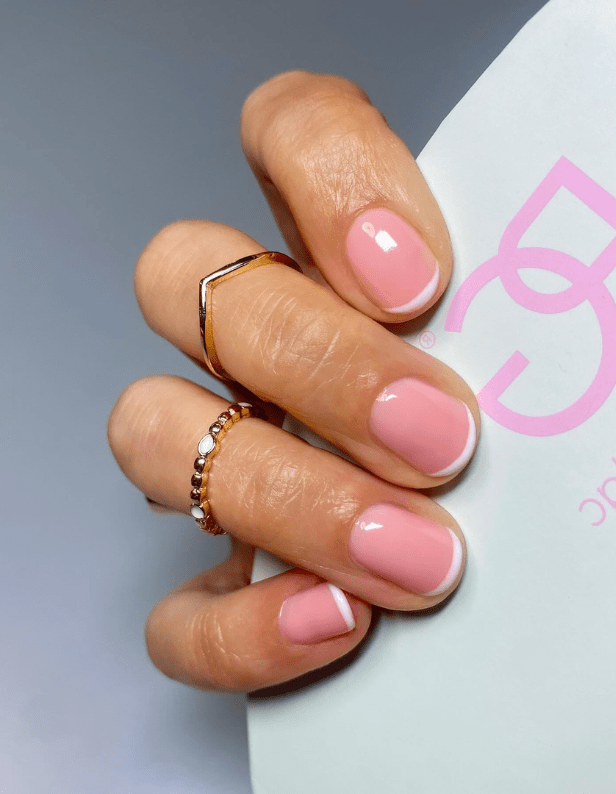 A hand holding a classic pink French manicure and two gold rings against a light-colored background.