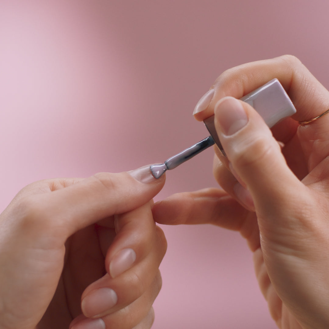 One hand applies silver-gray nail polish to another hand against a pink background.