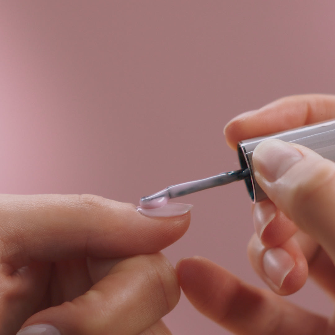Close-up of a person applying light pink nail polish to a nail with a brush, with a soft pink background.