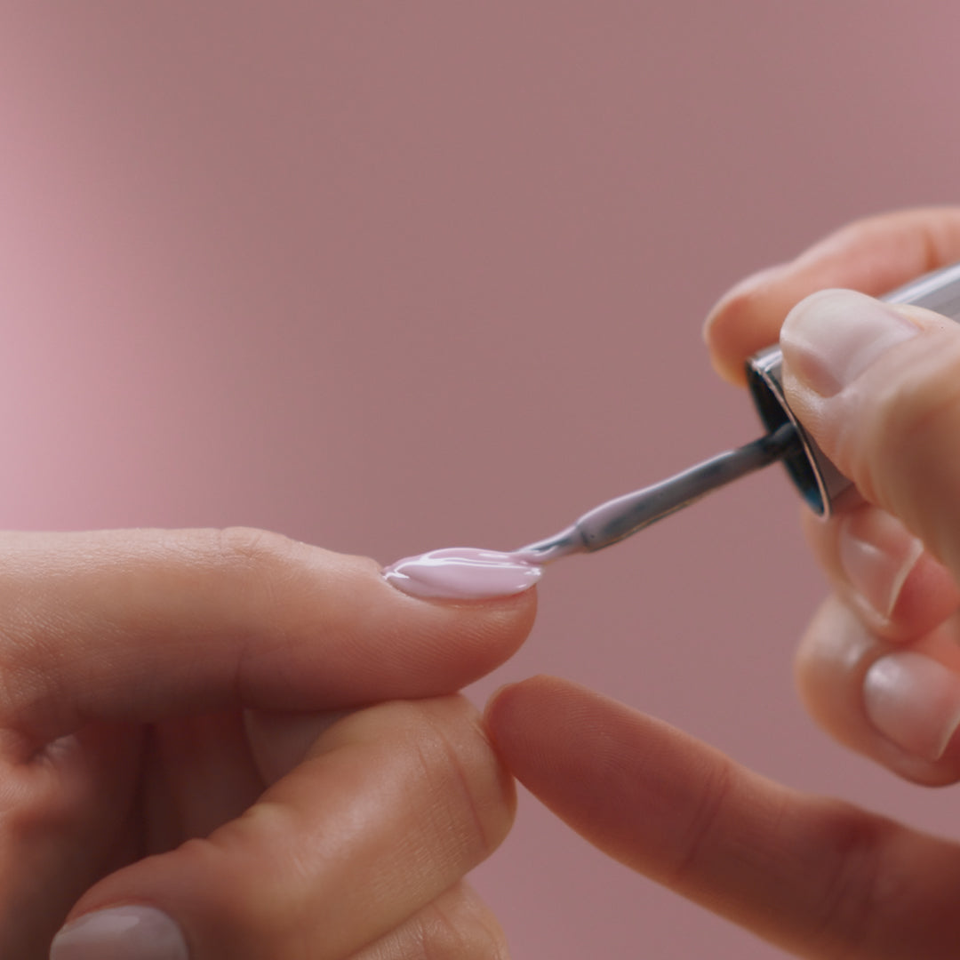 A close-up of a hand applying pale pink nail polish to a nail against a pink background.