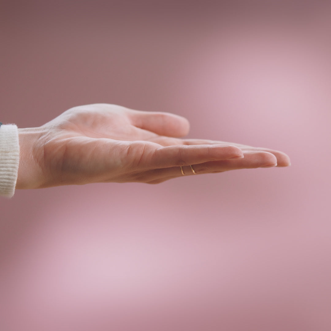 An open hand with gold rings, outstretched against a pink background.