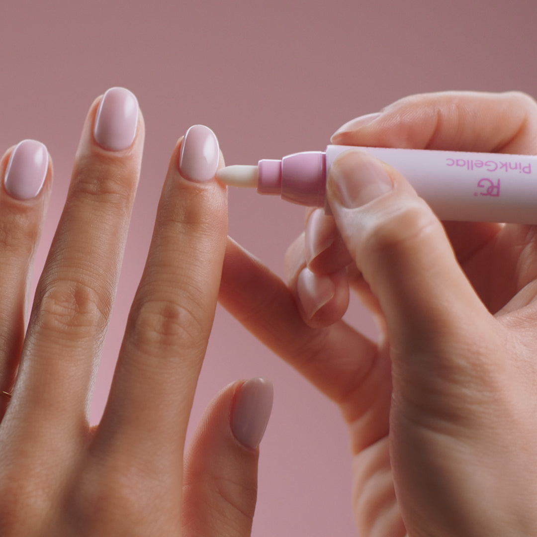 Two hands with well-groomed, light pink nails, with one©©hand using a cuticle pen on the cuticles of the other hand, against a pink background.