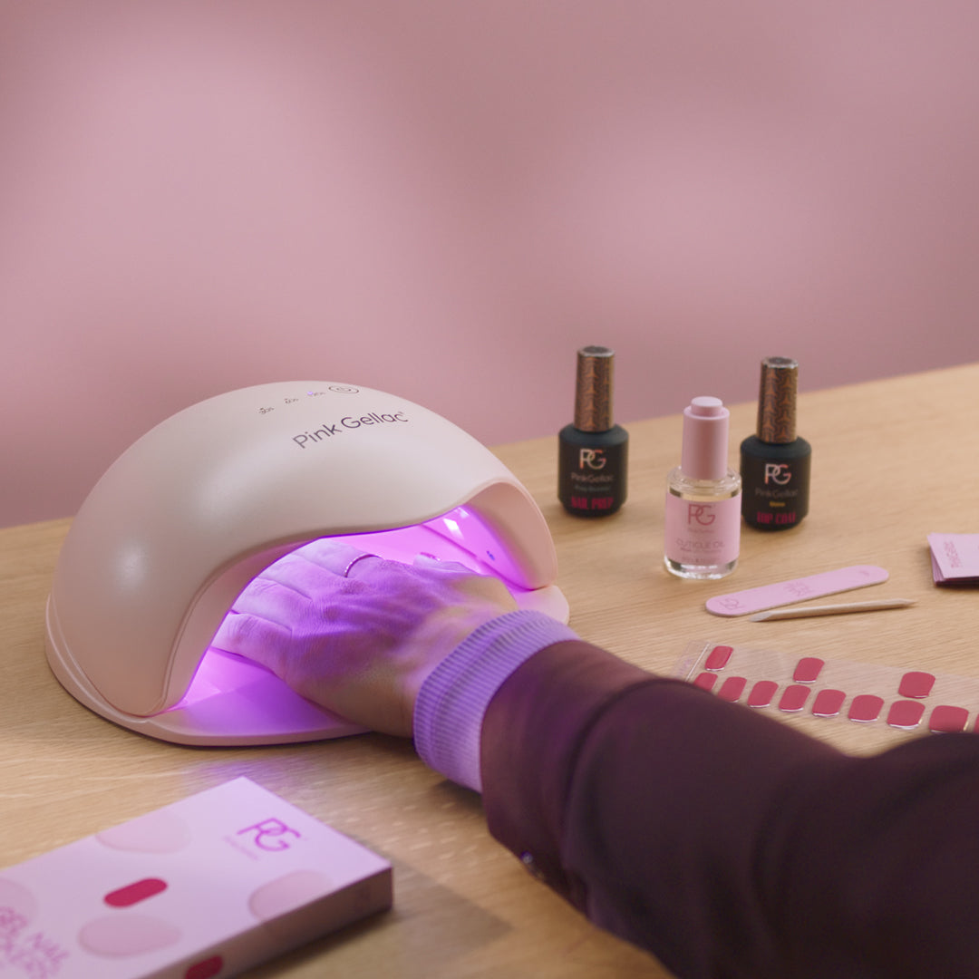 A hand under a UV lamp for nails with a few bottles of nail polish, a polishing file and color samples next to it on a wooden table.