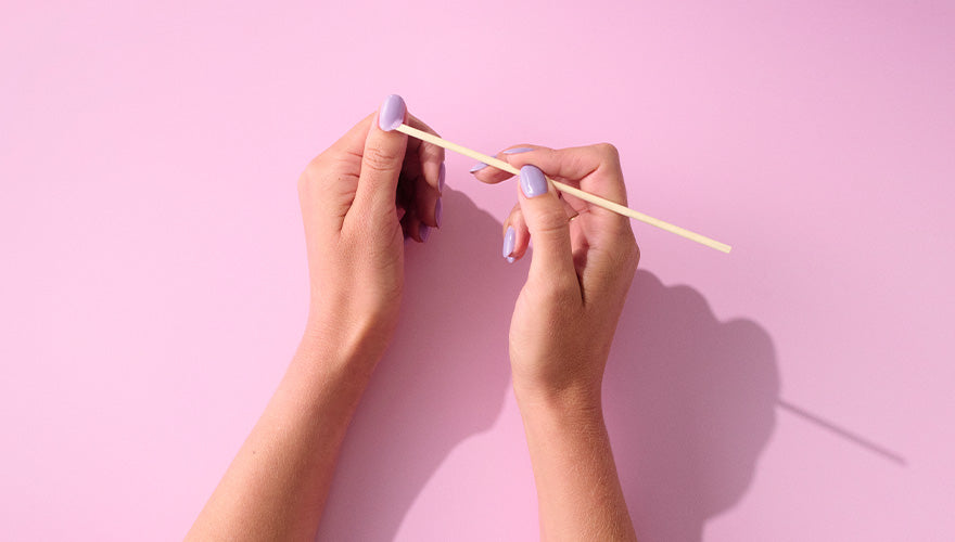 Two hands with purple nail polish hold a wooden stick against a pink background.