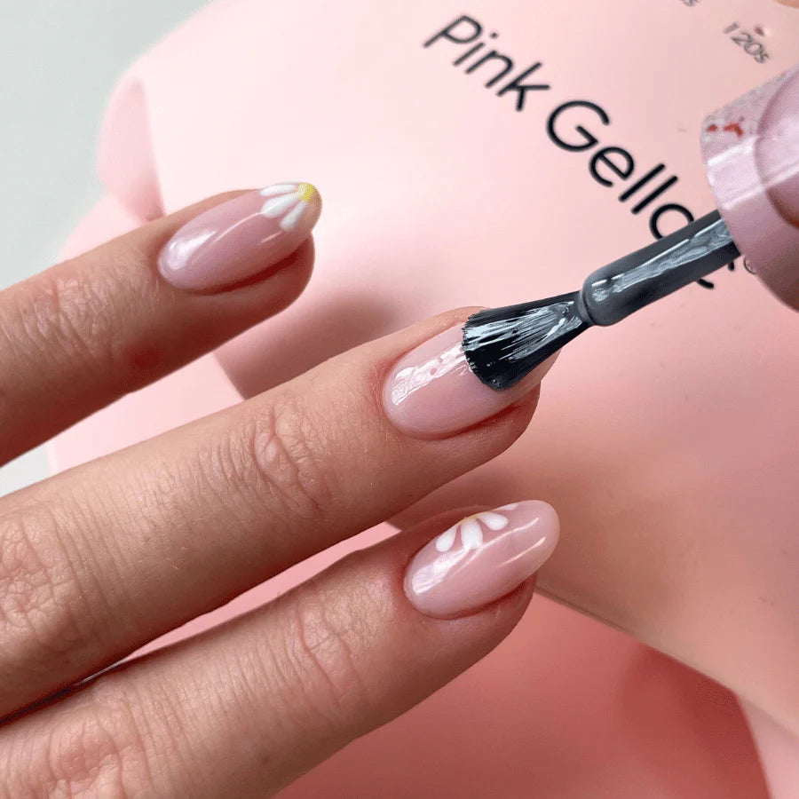 A hand with pink nail polish and floral design gets a shiny topcoat, with a pink packaging in the background.