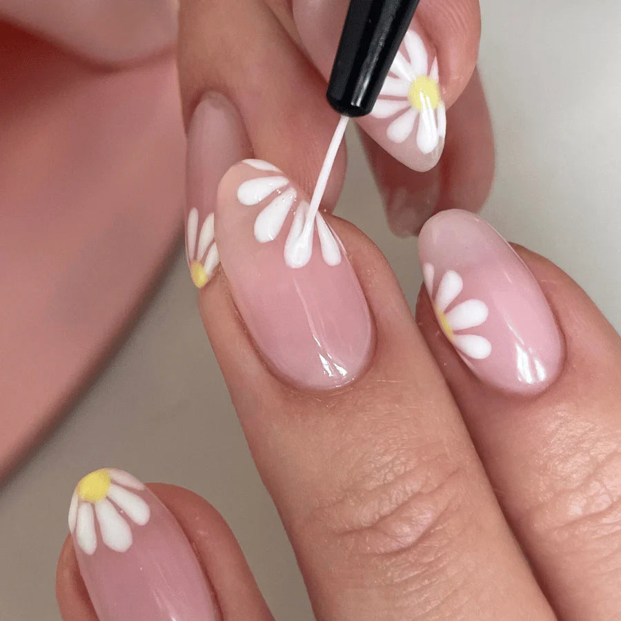 Close-up of a hand with a person painting white daisies on pink acrylic nails with a brush.