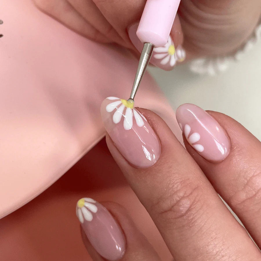 A hand with soft pink nail polish and white daisy nail art, using a dotting tool to create a flower on a nail.