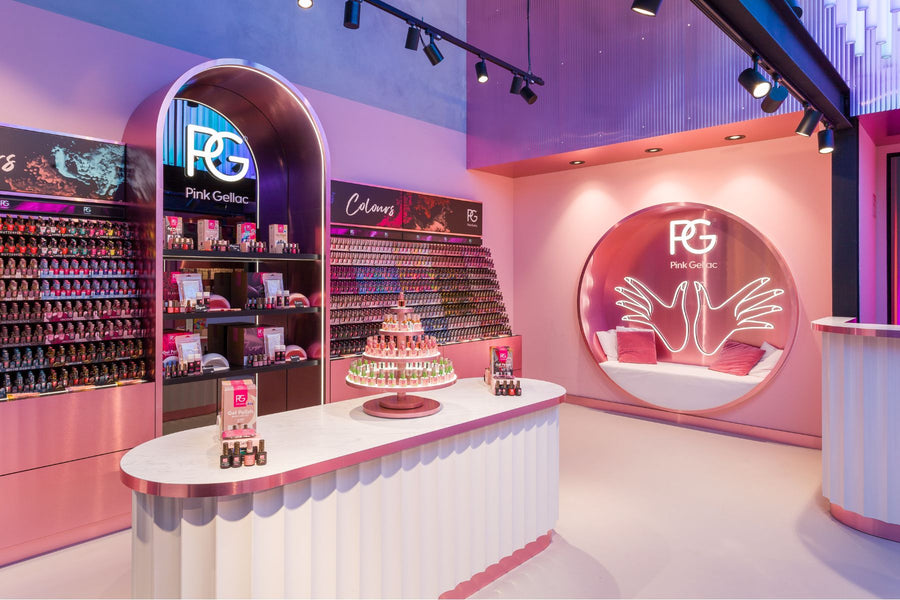 Interior of a modern pink-themed nail polish shop, with shelves full of coloured bottles of nail polish, a central display with a display window of products and a circular seating area with neon lighting in the shape of hands.