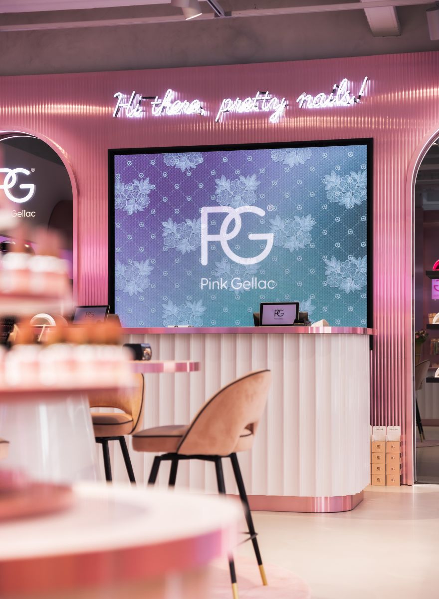 A stylish nail salon with a pink and white décor, soft chairs, a large screen with the Pink Gellac logo and the text "Hi there, pretty nails!" in neon lighting on the wall.