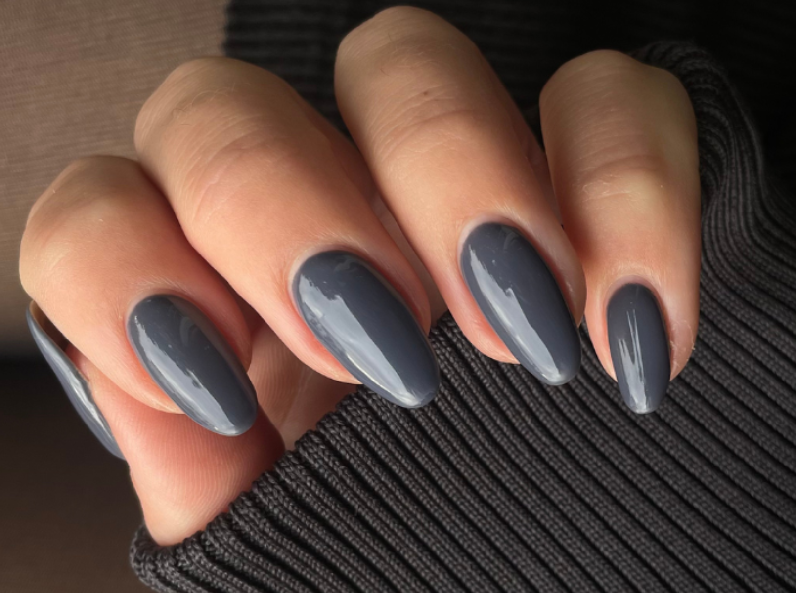 Close-up of a hand with long, oval nails painted in a shiny gray color, against a dark background of fabric.