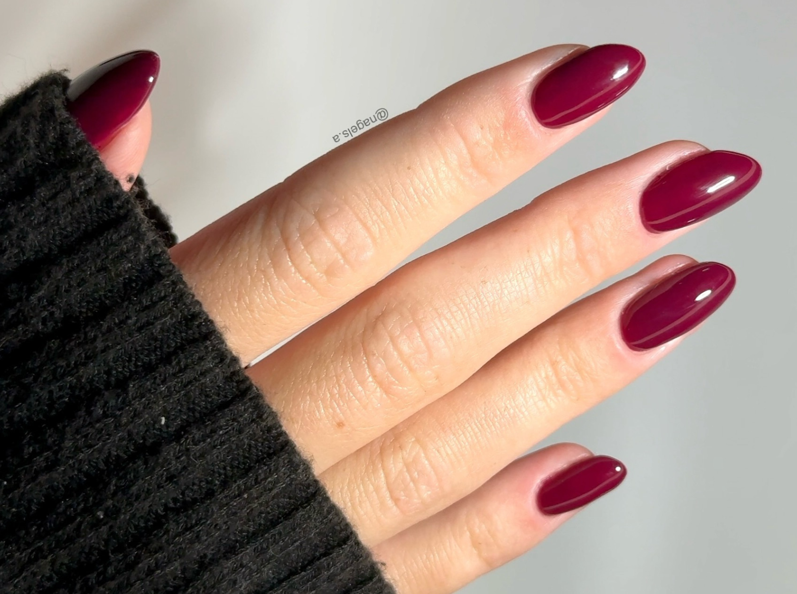 Close-up of a hand with burgundy, almond-shaped nails, wrapped in a black sweater.