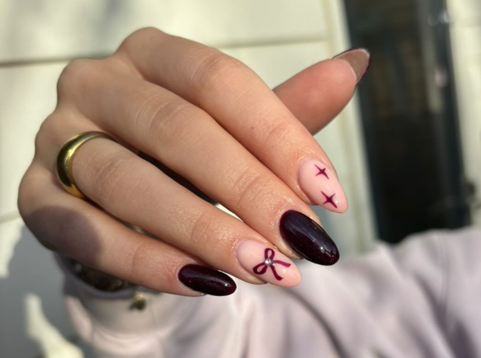 Hand with burgundy and pink nail designs, including bows and stars, and a gold ring on the middle finger.