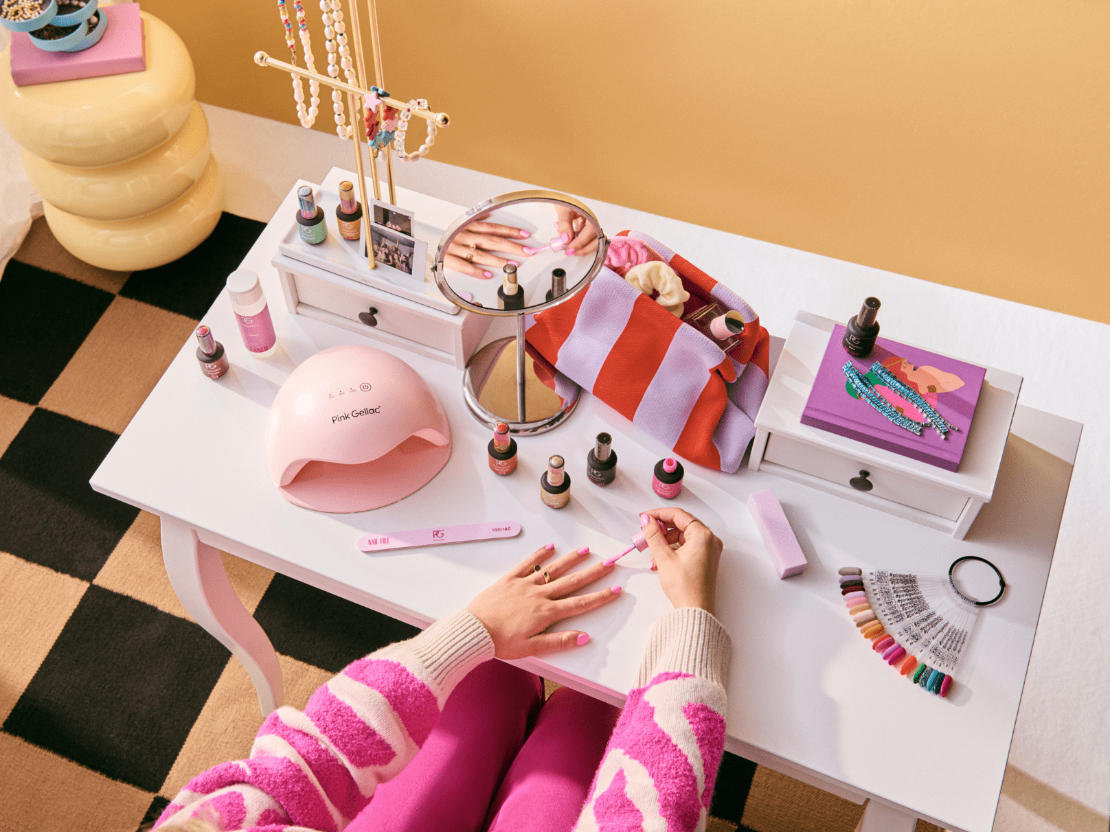 A person paints her nails at a white desk with several nail polish bottles, a pink nail dryer, a mirror, and jewelry stand on a striped purple and white bag.