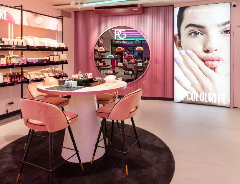 A beauty salon with pink chairs, a round table, a mirror with a logo above it, and a large poster of a woman with lilac nails on the wall.