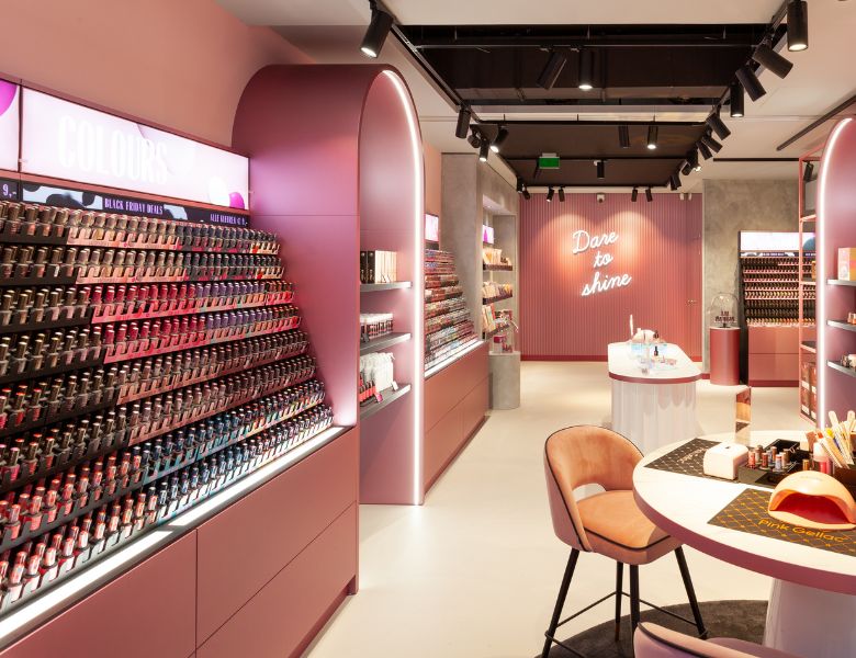 A colorful nail polish shop with well-stocked shelves, soft pink and purple tones, and a neon sign with the text "Dare to shine" in the background.