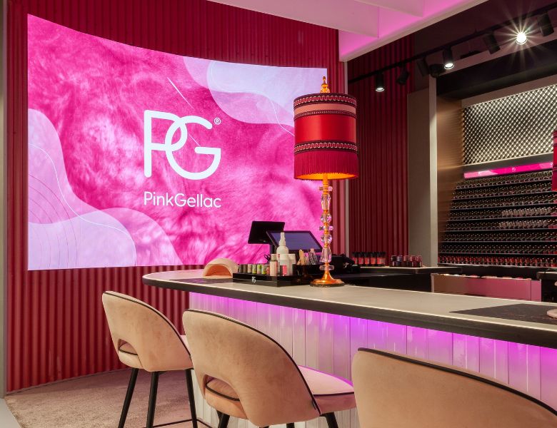 A modern nail salon with a large pink background with the logo "PG PinkGellac", a stylish lamp on the counter, and comfortable chairs.