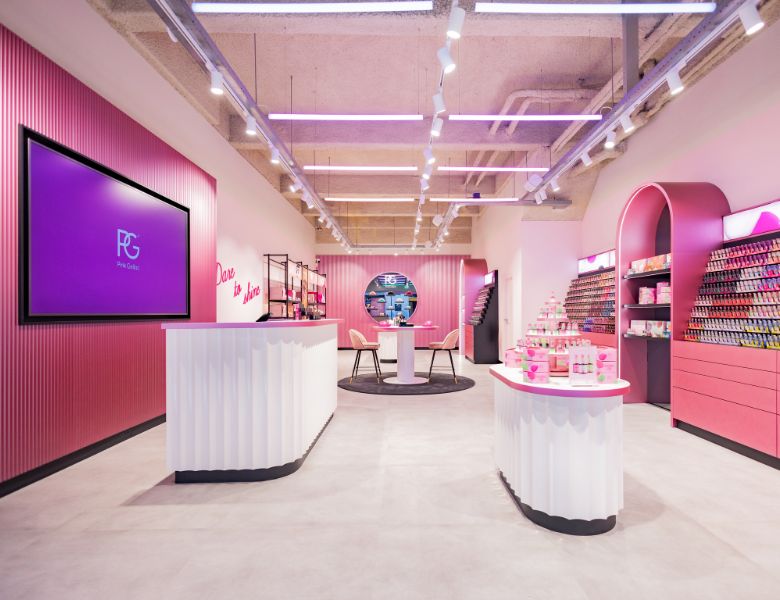 A modern store with pink and white décor, shelves filled with products, a round table with chairs, and bright pink lighting.