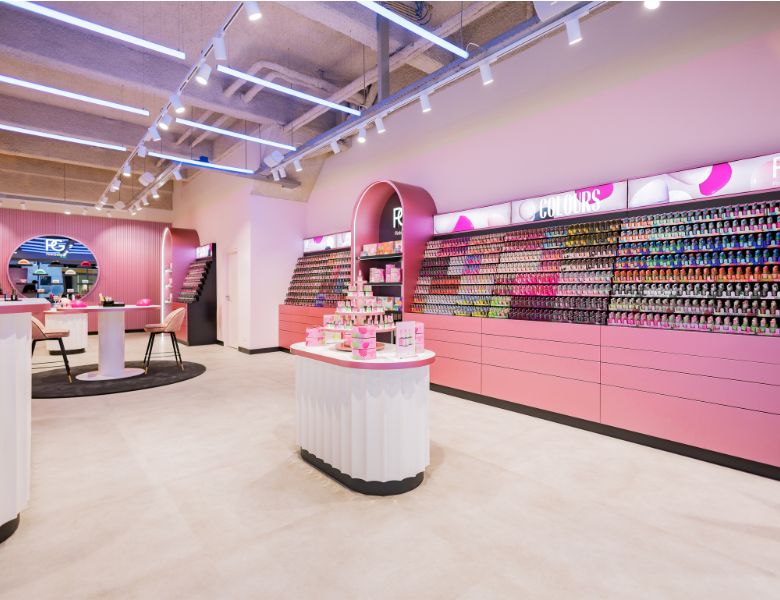 A modern store with pink décor, sleek lighting and a wall full of nail polish in various colors, with a central counter filled with product displays.