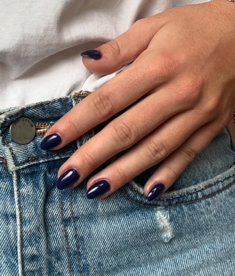 A hand with dark blue painted nails rests on a denim pocket.