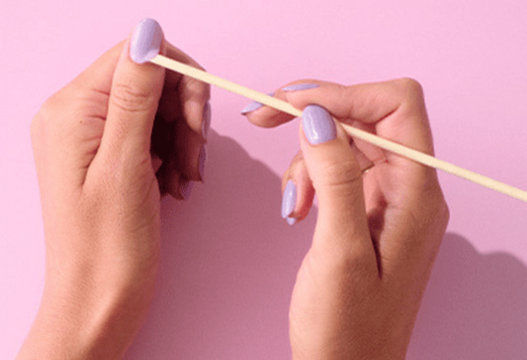 Two hands with lilac painted nails push back cuticles with a wooden cuticle stick on a pink background.