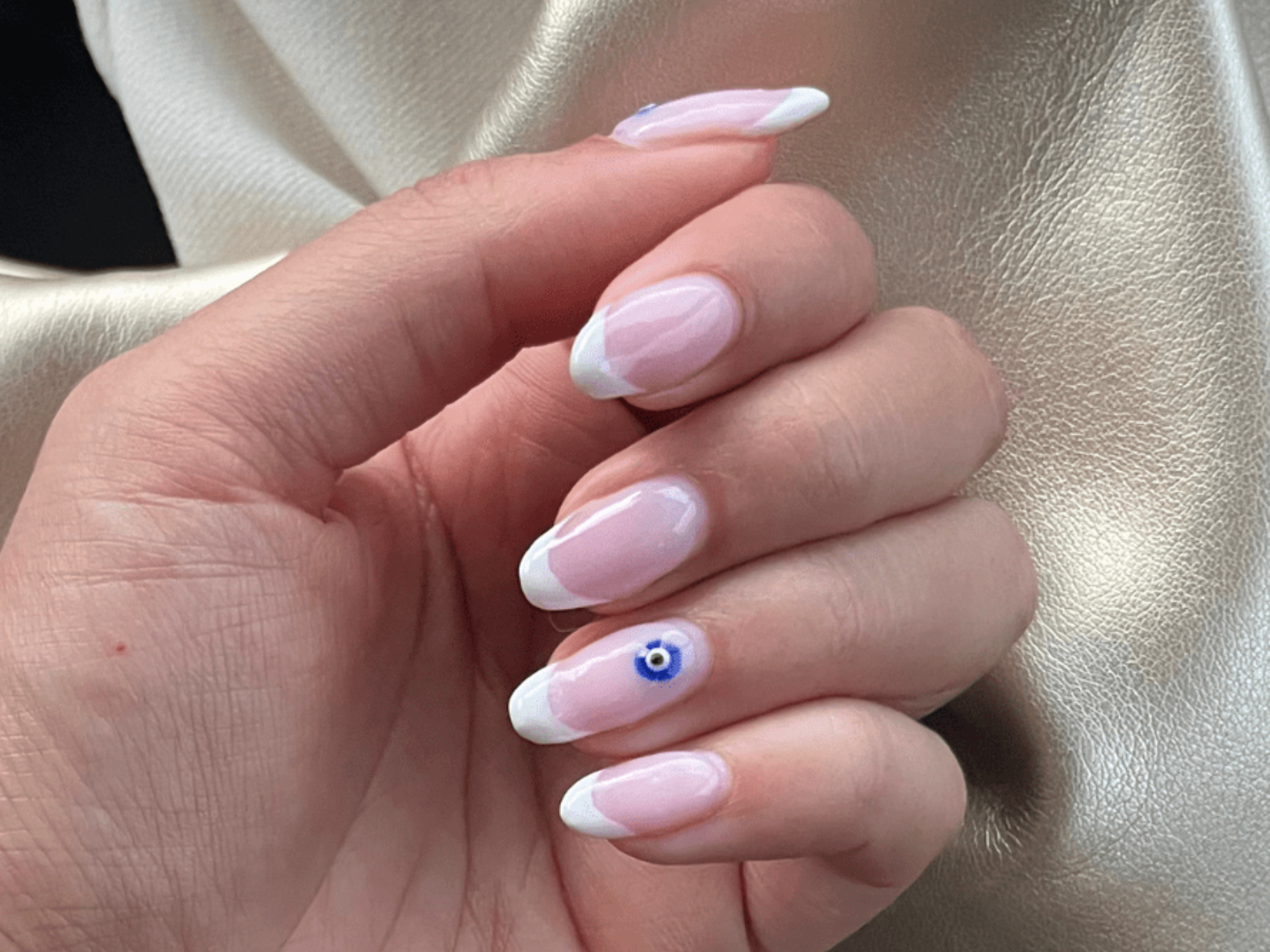 A hand with long acrylic nails with a French manicure and a blue eye design on the ring finger, against a shiny light background.