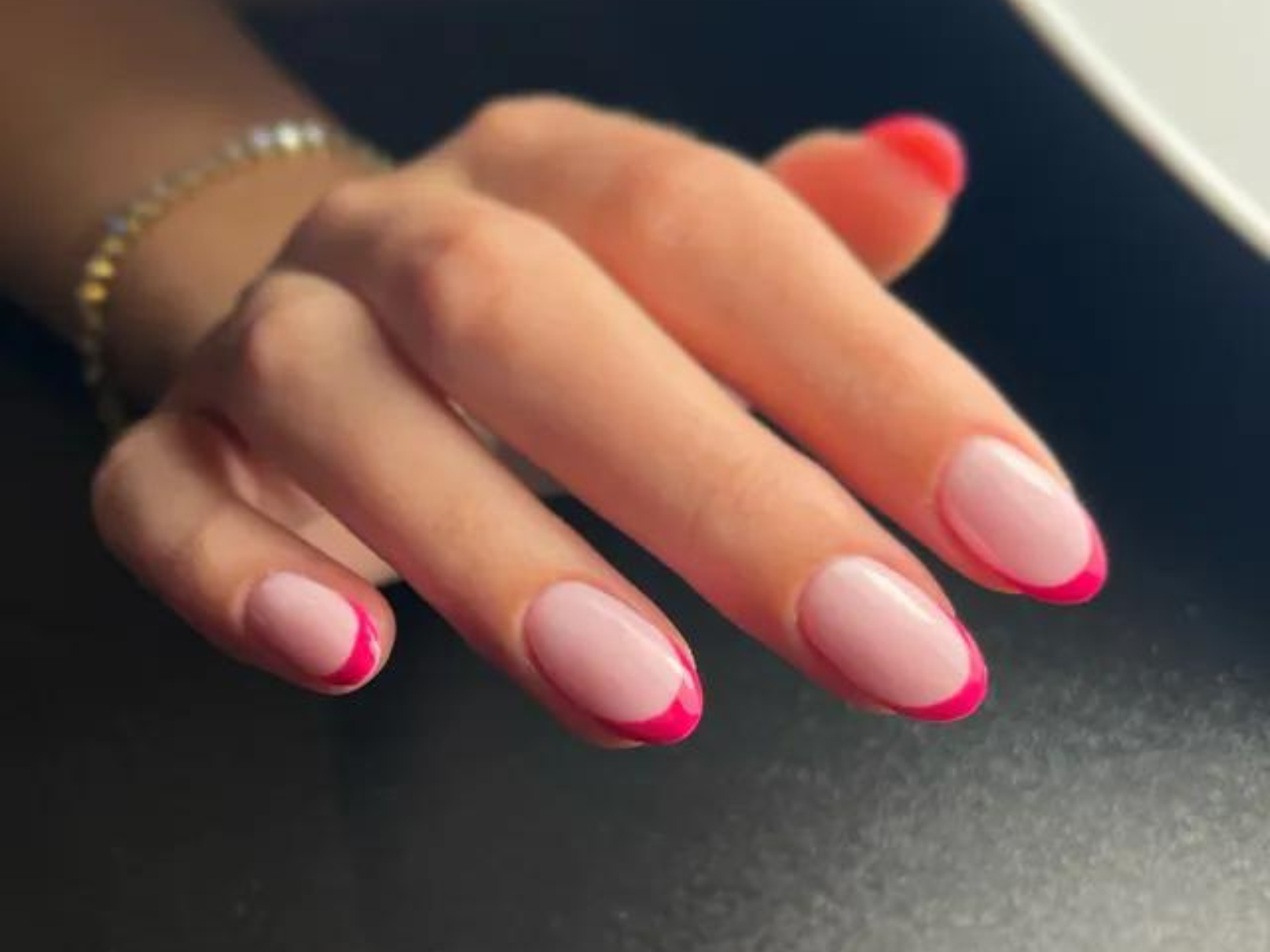 A hand with a bracelet and light pink nails with bright pink tips against a black background.