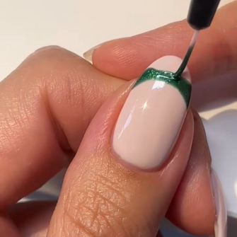 A hand using a nail polish brush to apply dark green nail polish to a beige painted nail.