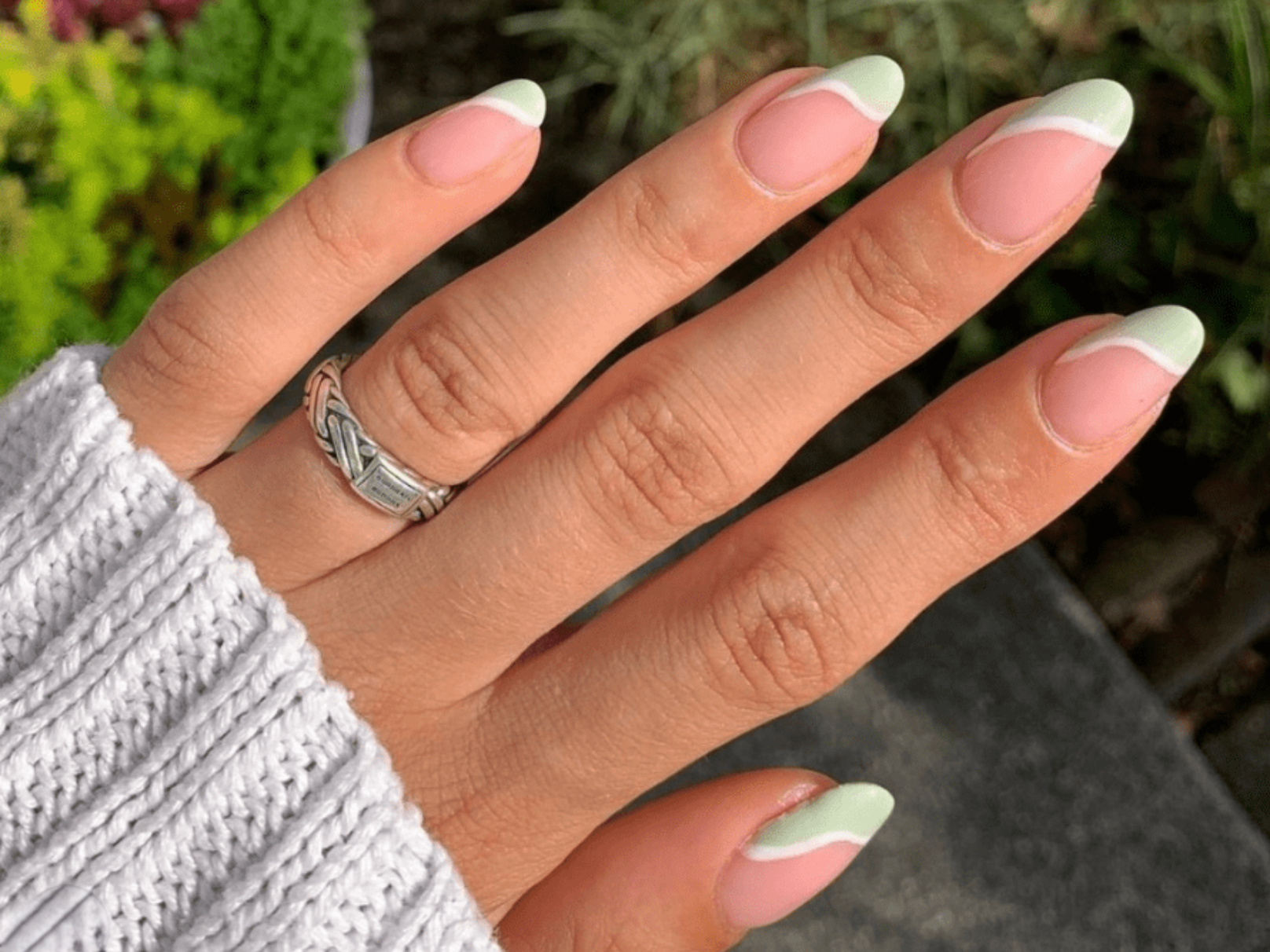 A hand dressed in a knitted sweater with long nails painted in a pastel green and pink design, and a silver ring on one of the fingers.