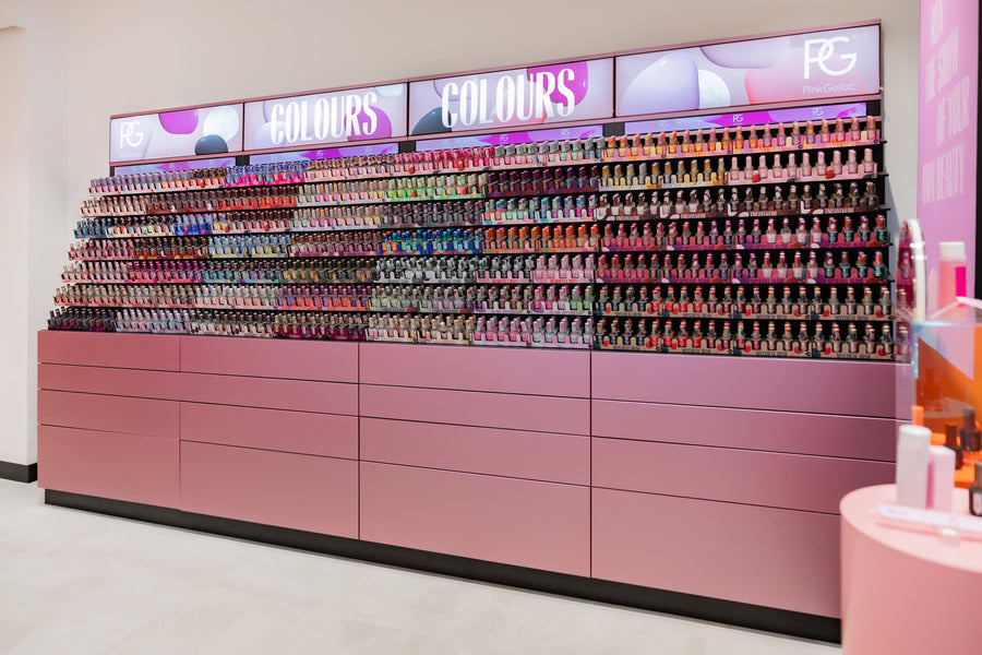 A wall with an elaborate display of colorful nail polish bottles, organized on pink shelves with illuminated advertising above them.
