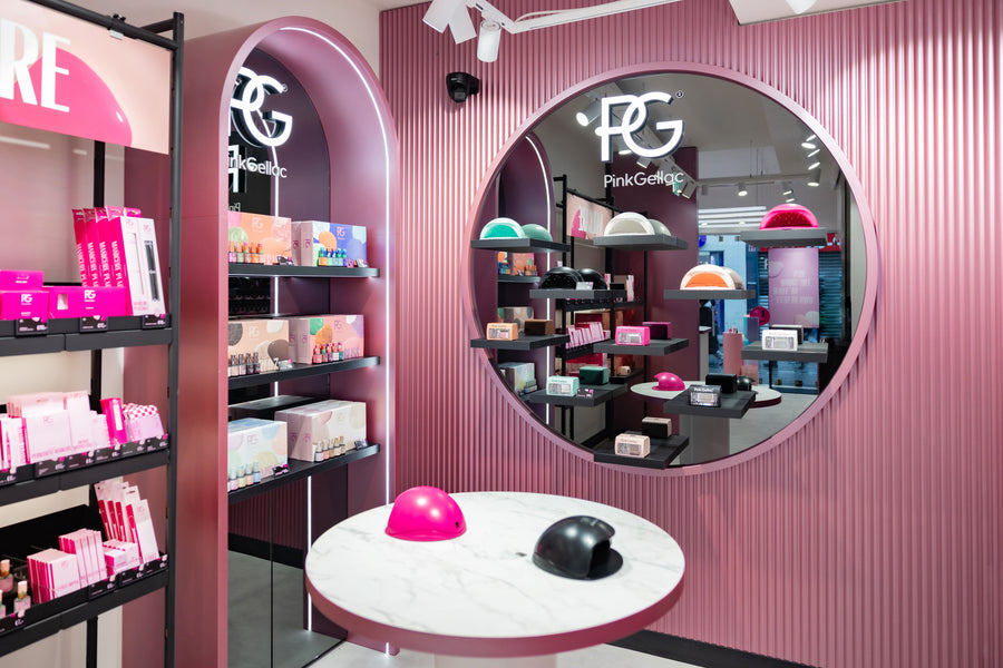 Pink shop interior with cosmetics products displayed on shelves, round mirror with logo and in the middle a table with a pink and black nail polish dryer.