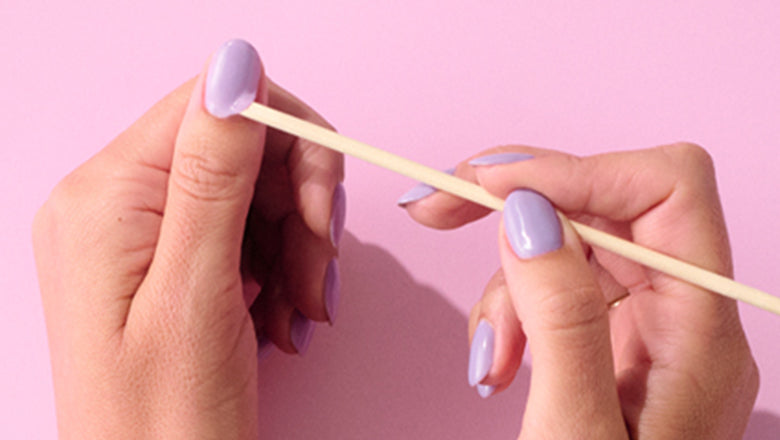 Two hands with lilac painted nails use a wooden stick against a pink background.