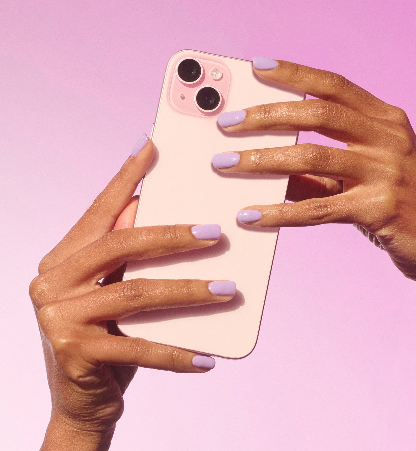 Two hands with lavender nails hold a pink smartphone against a pale pink background.