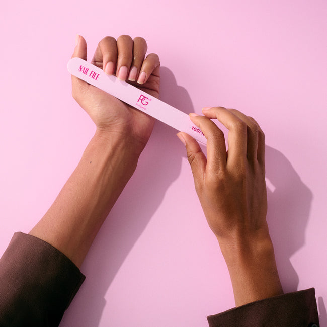 Two hands file nails with a pink nail file against a pink background.