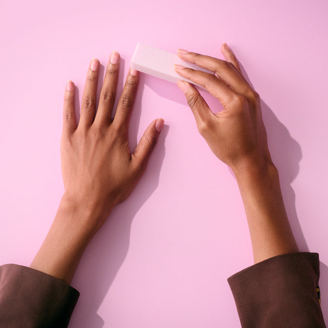 Two hands on a pink background, with one hand using a nail file on the nails of the other hand.