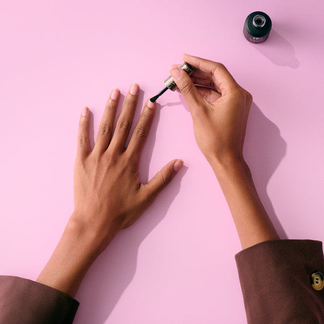 A person applying nail polish to her fingers on a pink background.