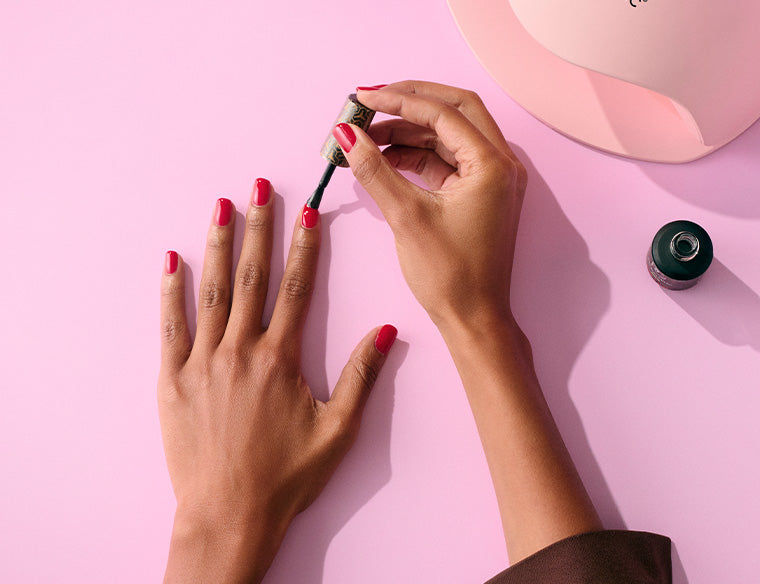 Hand with red nail polish painting nails on a pink background, with an open jar of nail polish and a UV lamp visible.