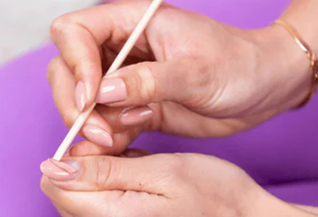 Hands editing cuticles with wooden stick, pink painted nails, purple background.