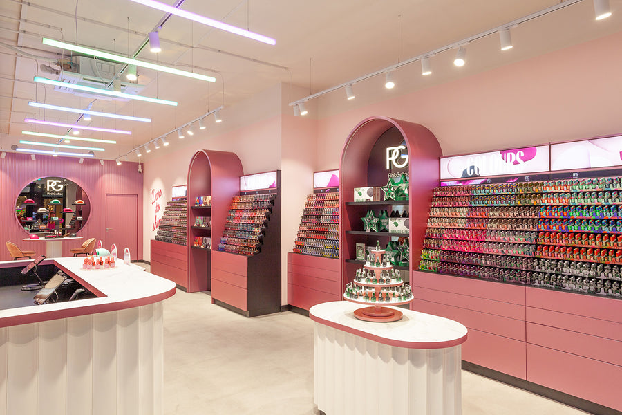 A modern nail salon with pink and purple accents, illuminated shelves full of nail polish in different colors, a round mirror on the wall and a central counter.