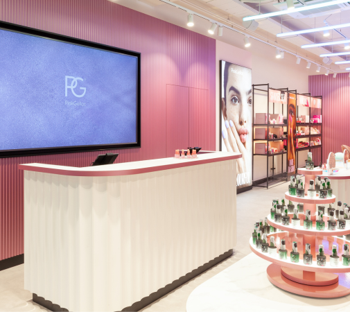 A modern and stylish store interior with a pink color scheme, a white reception desk, a large screen with the letters "PG", and a display of cosmetics and skincare products on circular shelves.