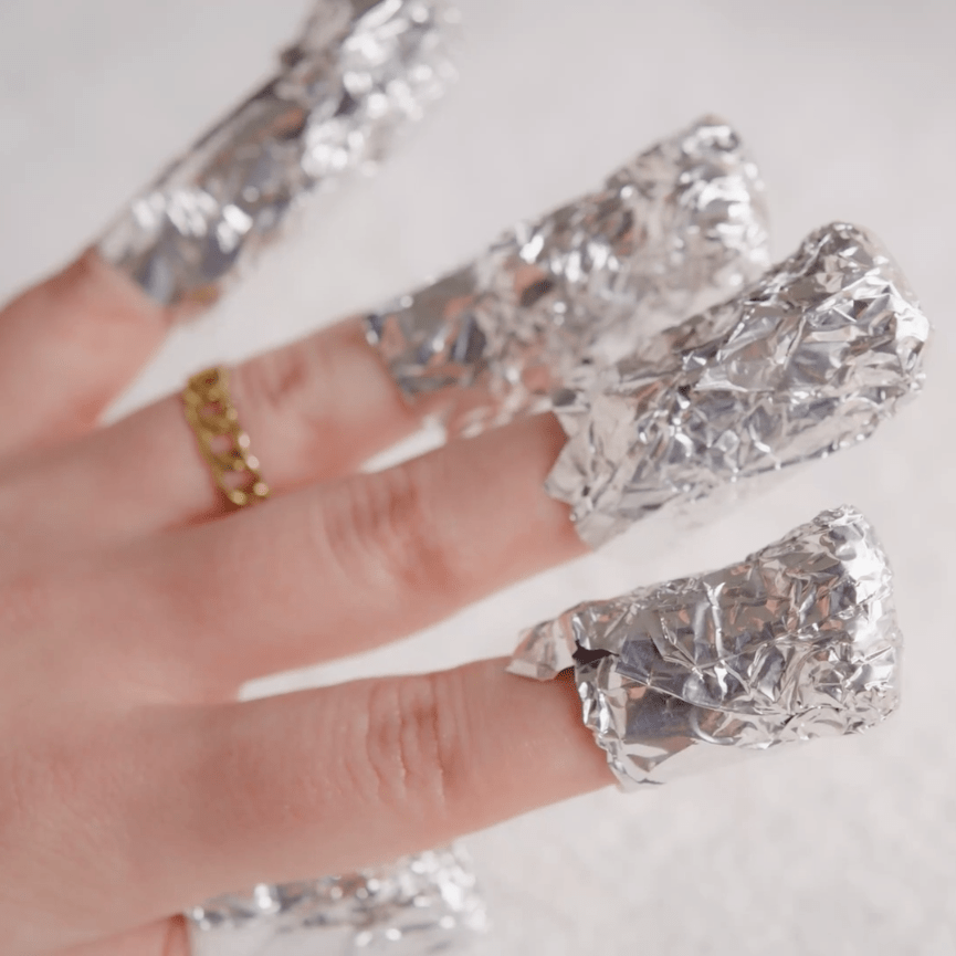 Fingers wrapped around the nails with aluminum foil for nail polish removal, with a gold ring on the middle finger.