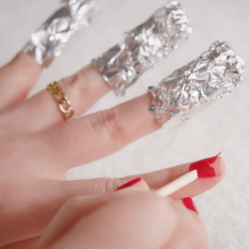 Person with aluminum foil on three fingers and red nail polish on one©©nail, removing nail polish with a wooden stick.