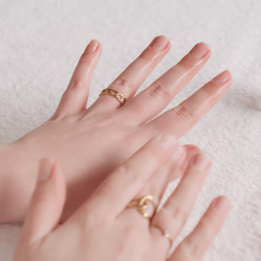 Two hands with gold-colored rings are massaged with oil on a white towel.