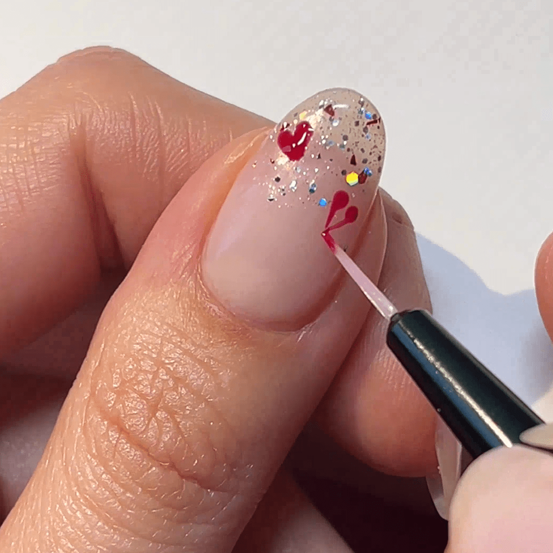 Nail art with red hearts and glitter is applied on a transparent nail.